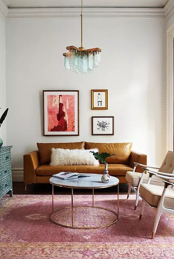 a colorful living room with a rust colored leather sofa, beige chairs, a round table, a pink rug and a blue inlay dresser, a whimsical mini gallery wall