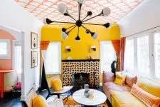 a colorful living room with a yellow accent wlal, a bold wallpaper ceiling, a pink and yellow sofa, rattan chairs and yellow pillows