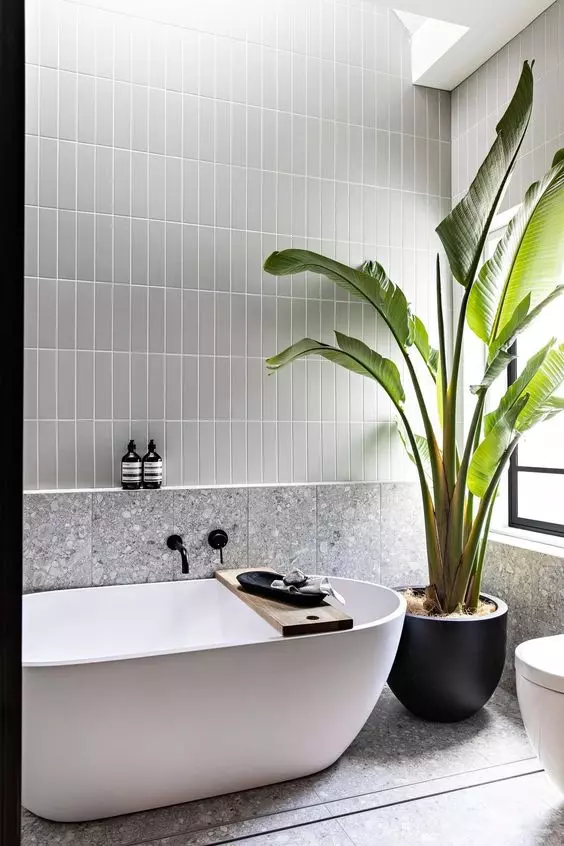 a contemporary bathroom with grey stacked tiles and grey terrazzo ones, an oval tub, a potted plant and some black decor pieces