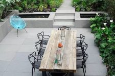 a contemporary garden with lots of greenery and blooms, clad with concrete tiles, a wooden table and black chairs