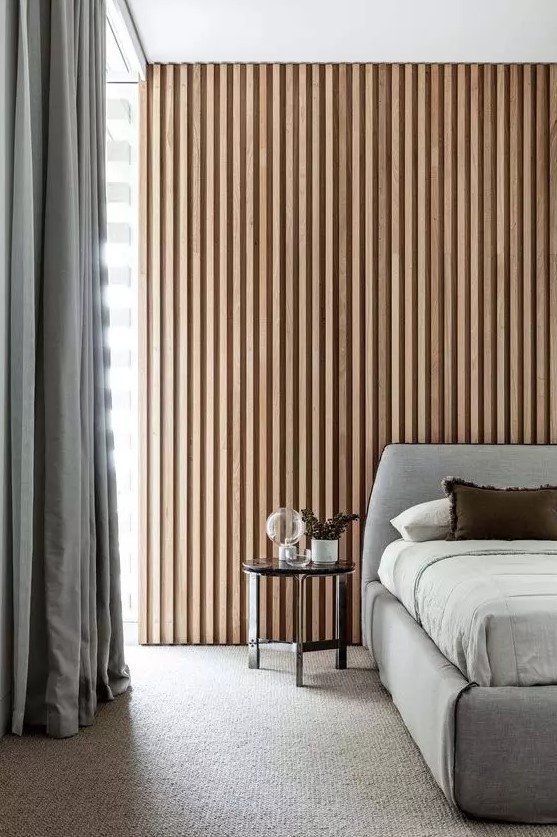 a contemporary neutral bedroom with a wood slat accent wall, a grey upholstered bed with neutral bedding, grey curtains and a black side table