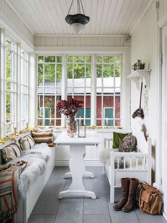 a cozy Scandinavian sunroom with a built-in bench, a white table, a usual bench, lots of pillows and blankets, greenery and foliage