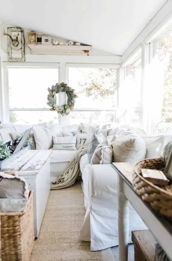 a cozy neutral farmhouse sunroom with white furniture, baskets, a shabby chic table, a leaf wreath and white pumpkins