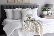 a farmhouse bedroom with grey walls, a grey upholstered bed with white bedding, a grid gallery wall of vintage herbs and blooms