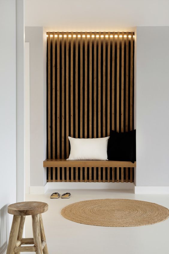 a laconic Japandi entryway with a wood slat accent wall and lights, a built-in daybed and a wooden stool and jute rug
