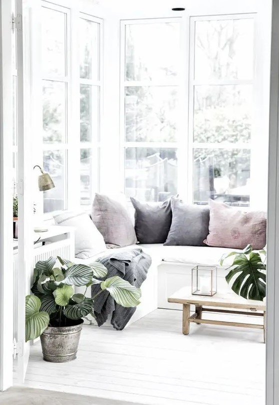 a lovely Nordic sunroom in white, with a built-in sectional sofa with pastel pillows, potted plants and a low wooden coffee table