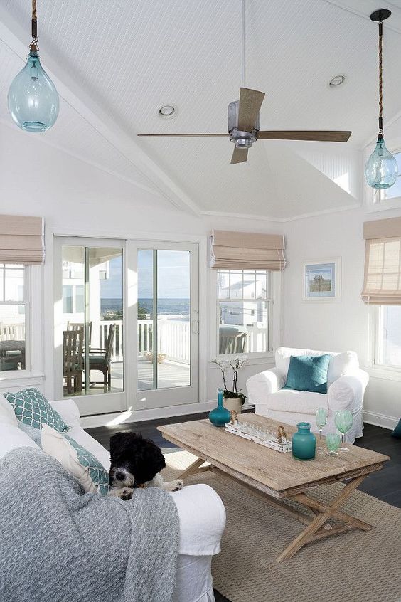 a lovely coastal sunroom with white seating furniture, blue and aqua pillows and blankets, a stained table and blue vases and lamps