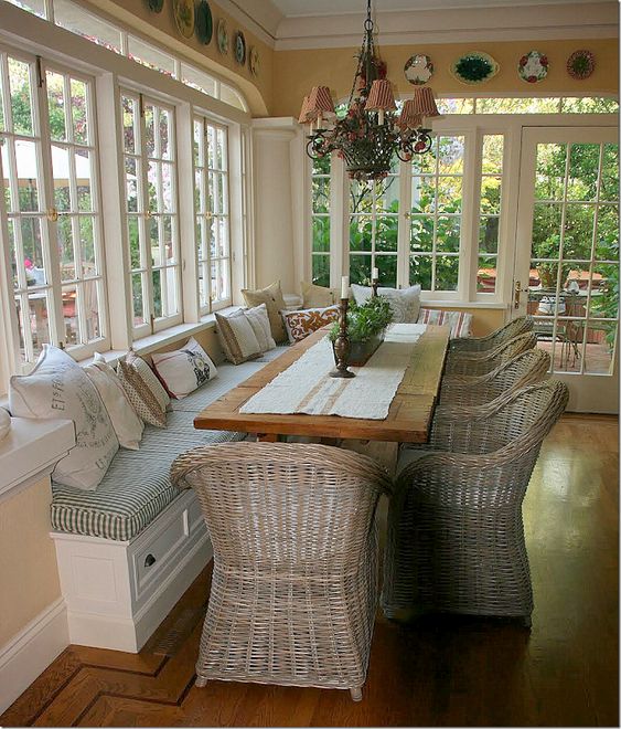 a lovely famrhouse sunroom with a built-in bench, a stained table and wicker chairs, a vintage chandelier and a gallery wall of plates