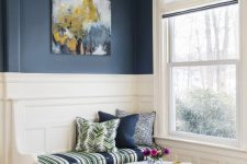 a lovely reading nook by the window, with navy walls, white paneling and wallpaper o the ceiling, a built-in bench and a round pouf