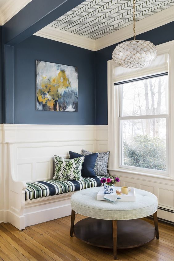 a lovely reading nook by the window, with navy walls, white paneling and wallpaper o the ceiling, a built in bench and a round pouf