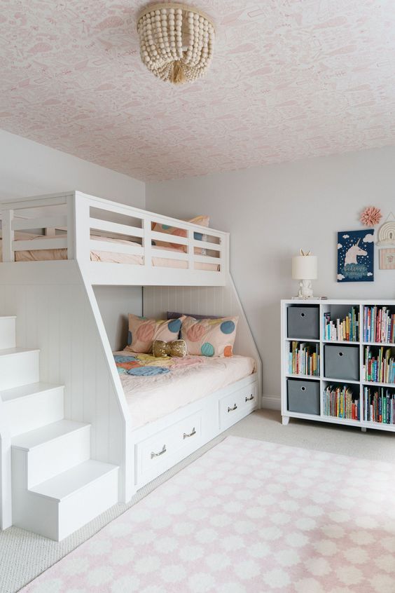 a lovely shared girls' bedroom with a pink wallpaper ceiling, a white bunk bed with bold bedding, a storage unit and some art