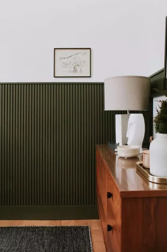 a mid-century modern living room with dark green wood slat panels on the wall, a stained dresser, a table lamp and some greenery