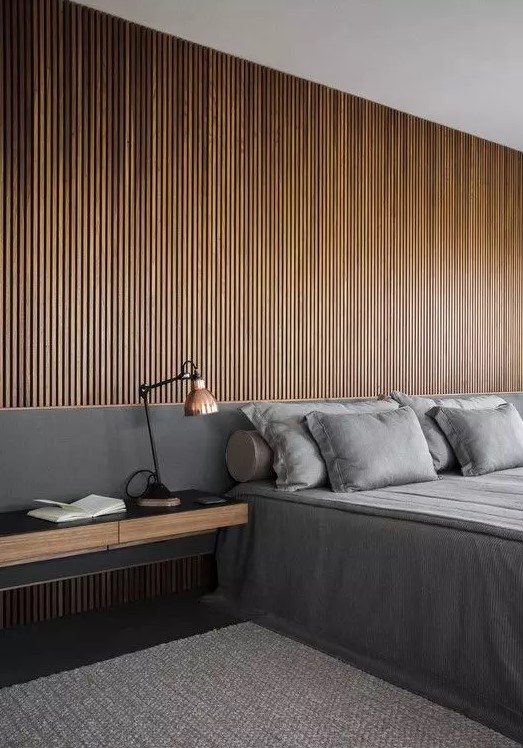 a minimalist meets contemporary bedroom with a wood slat accent wlal, a grey bed with grey bedding, a floating nightstand and a table lamp