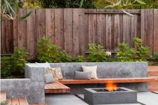 a minimalist outdoor space with a concrete and wood built-in bench and a concrete fire pit, with greenery around welcomes in