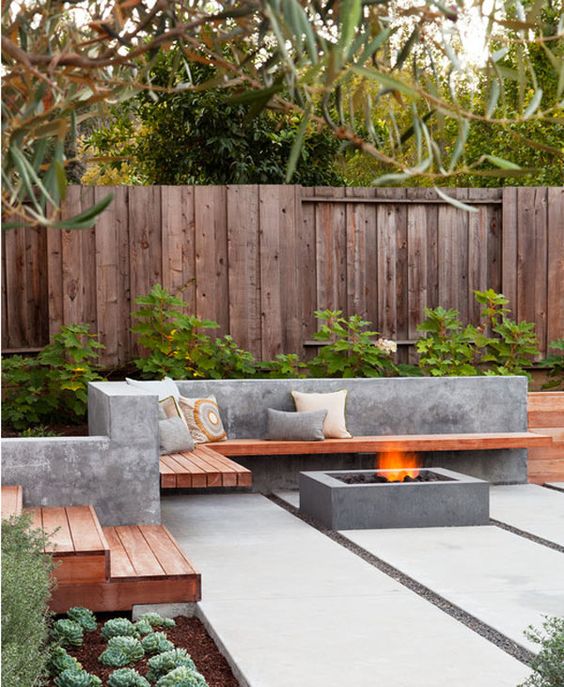 a minimalist outdoor space with a concrete and wood built-in bench and a concrete fire pit, with greenery around welcomes in