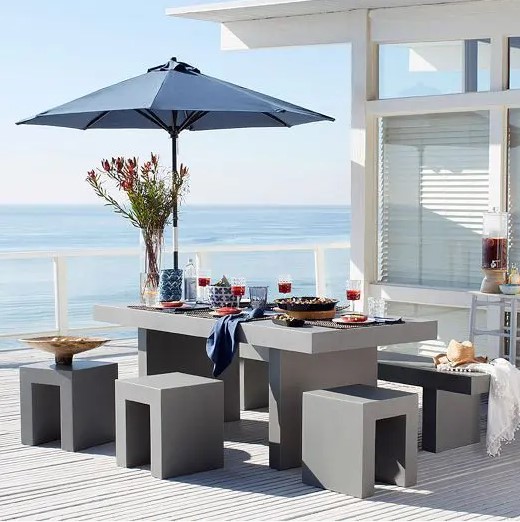 a minimalist seaside terrace with a concrete table and stools, a navy umbrella and some blankets plus an amazing sea view