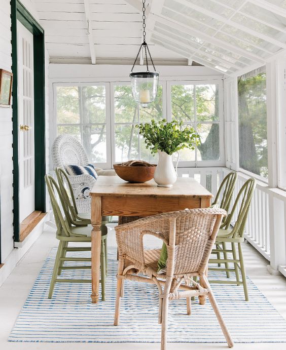 a modern farmhouse sunroom with a stained table, green wood and wicker chairs, a candle pendant lamp and some greenery