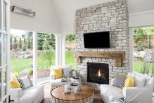 a modern farmhouse sunroom with white furniture, a stone clad fireplace, a rattan table and sunny yellow accents