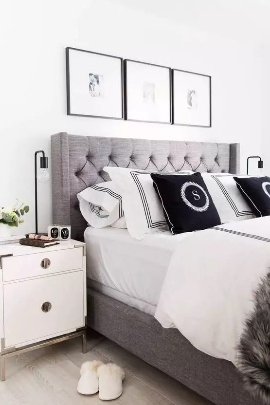 a modern glam bedroom with a grey upholstered bed with black and white bedding, a neutral nightstand with brass legs and a grid gallery wall