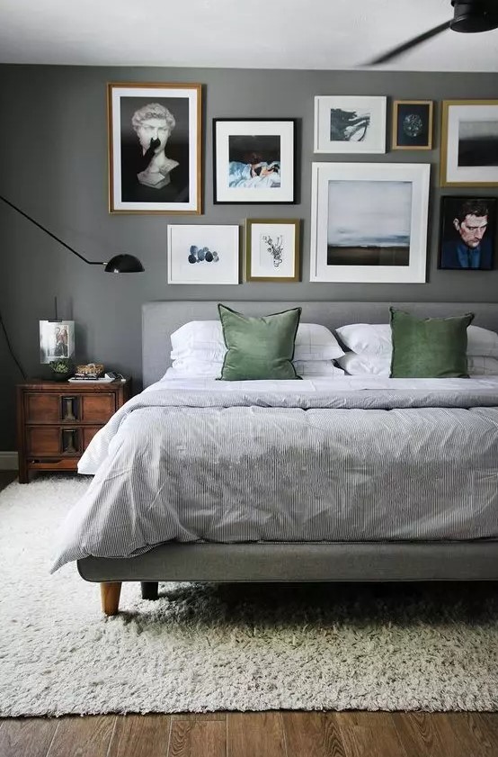 a moody grey bedroom with a grey upholstered bed with grey and green bedding, stained nightstands, a free form gallery wall