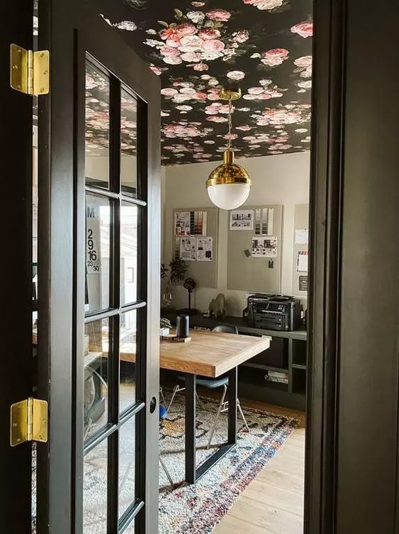 a moody home office with a black floral wallpaper ceiling, a chic desk, a grey storage unit, a printed rug and some memo boards