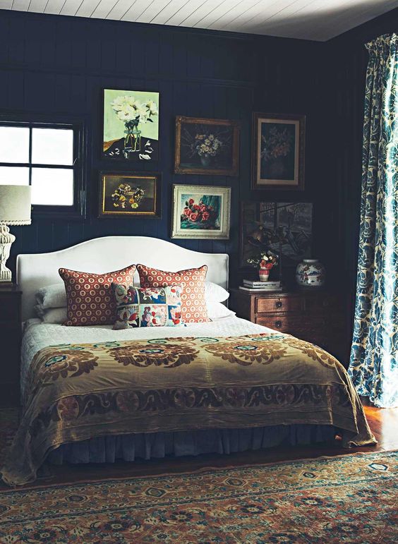 a moody vintage bedroom with a white upholstered bed, stained nightstands, a vintage artwork gallery wall and prined textiles