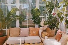 a neutral boho sunroom with a pallet corner sofa, bright pillows, a wicker coffee table, potted plants and pampas grass
