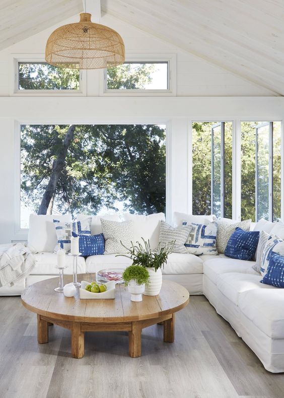 a neutral coastal sunroom with a white corner sofa with printed pillows, a round stained table and a wicker lamp