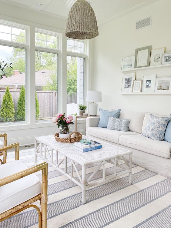 a neutral coastal sunroom with neutral furniture, a low coffee table, a woven pendant lamp, printed textiles and a gallery wall
