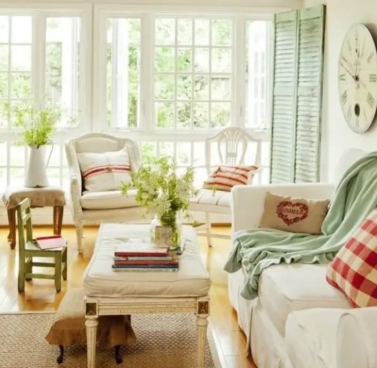 a neutral cozy farmhouse sunroom with white vintage furniture, green shutters and blankets, touches of plaid and stripes plus greenery