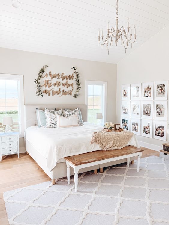 a neutral farmhouse bedroom with tan walls, a tan upholstered bed, a wooden bench, white nightstands and a grid gallery wall of family pics