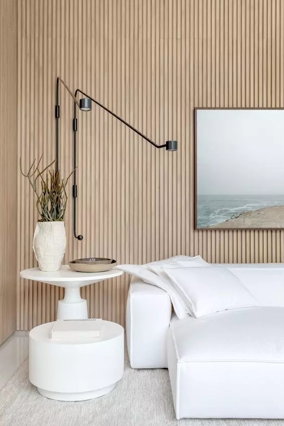 a neutral living room with a wood slat accent wall, a white sofa and pillows, white coffee tables and a scenery is a beautiful modenr interpretation of a coastal interior