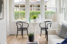 a pretty Scandinavian sunroom with a creamy sofa with printed pillows, a white round table, black chairs, a wooden bench and a wooden chair