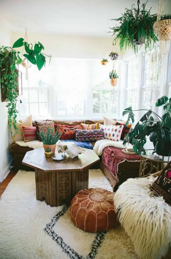 a pretty and bright boho sunroom with a sectional, colorful pillows and blankets, a carved wooden table, a leather pouf and potted plants