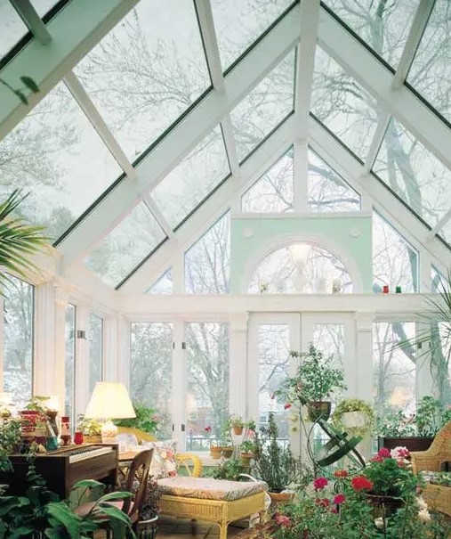 a refined and chic sunroom with a piano, rattan furniture, lots of potted blooms and greenery and lamps
