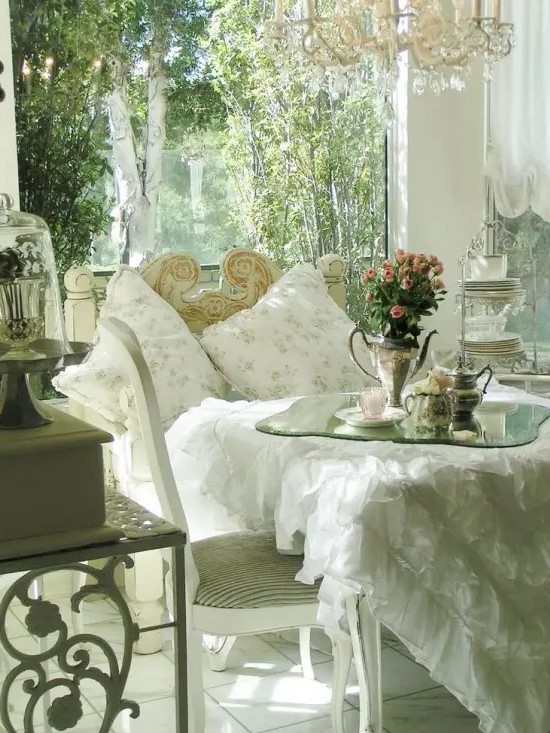 a refined white vintage sunroom with shabby chic and vintage furniture, a crystal chandelier, floral pillows and blooms in vases
