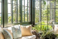 a rustic sunroom with green window frames, wicker corner sofas, a stained coffee table and potted plants plus amazing forest views