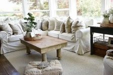 a simple farmhouse sunroom with an L-shaped white sofa, potted greenery, a rustic table and neutral textiles