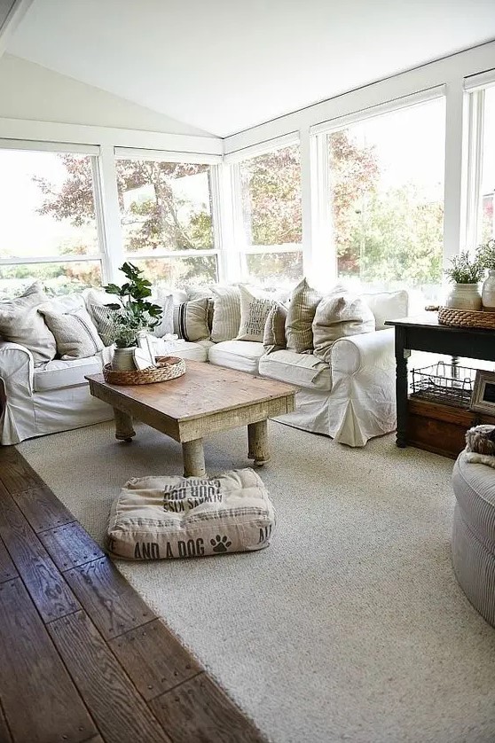 a simple farmhouse sunroom with an L-shaped white sofa, potted greenery, a rustic table and neutral textiles