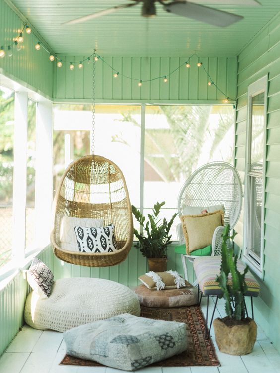 a small and cozy boho sunroom with green walls, a series of windows, a pendant chair, a papasan chair, some cushions and pillows and a bold rug