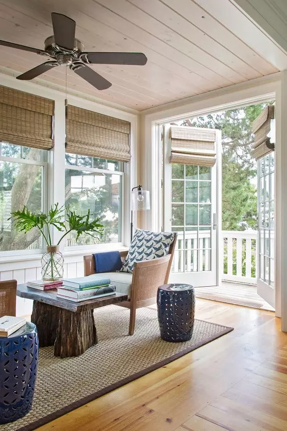 a small and welcoming sunroom with neutral chairs, a rough wood table, navy side tables and woven blinds