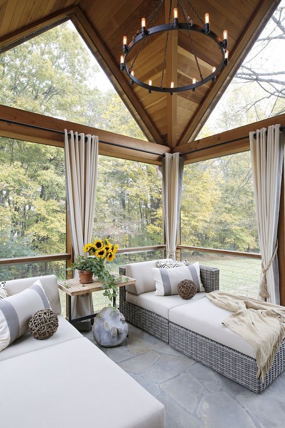 a small rustic sunroom with extensive glazing, wicker daybeds, a side table, a vintage chandelier and lots of pillows