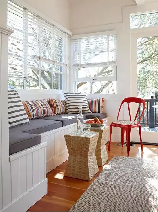 a small sunroom living room with a large built in bench with storage and a duo of wicker stools
