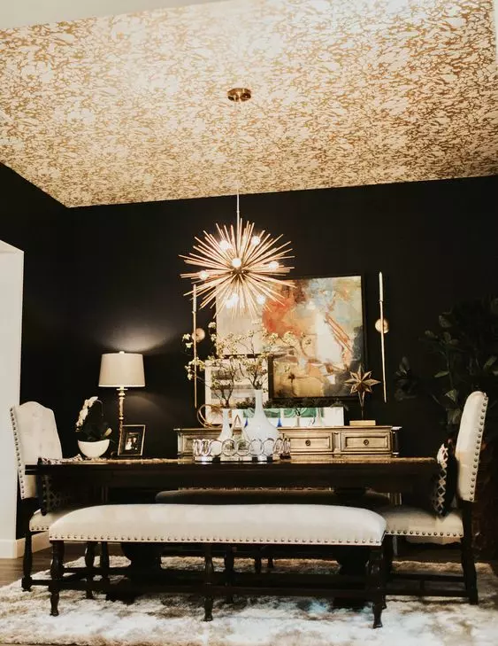 a sophisticated dining room with black walls, a gold wallpaper ceiling, a large table, creamy chairs and a bench and a credenza