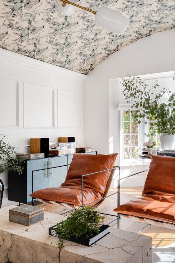 a sophisticated living room with an arched wallpaper ceiling, white paneling, a dark green credenza, amber leather chairs and a slab coffee table
