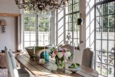 a sophisticated vintage sunroom with a series of French windows, a unique dining table with a planked top, neutral chairs and a lovely crystal chandelier