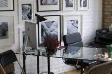 a stylish home office with a white brick accent wall with a gallery wall, a glass desk, black leather chairs and some potted plants