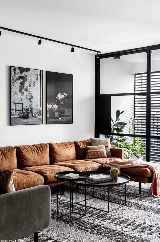 a stylish living room with a rust sectional, a grey chair, black round tables, a black and white gallery wall and some potted plants