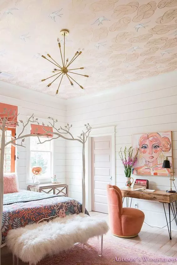 a subtle bedroom with a printed wallpaper ceiling, a bed with printed bedding, a bench with white fur, coral textiles and a chair