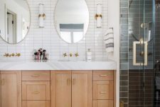 a two-tone bathroom with white and black stacked tiles, a light-stained vanity, oval mirrors, brass fixtures and sconces